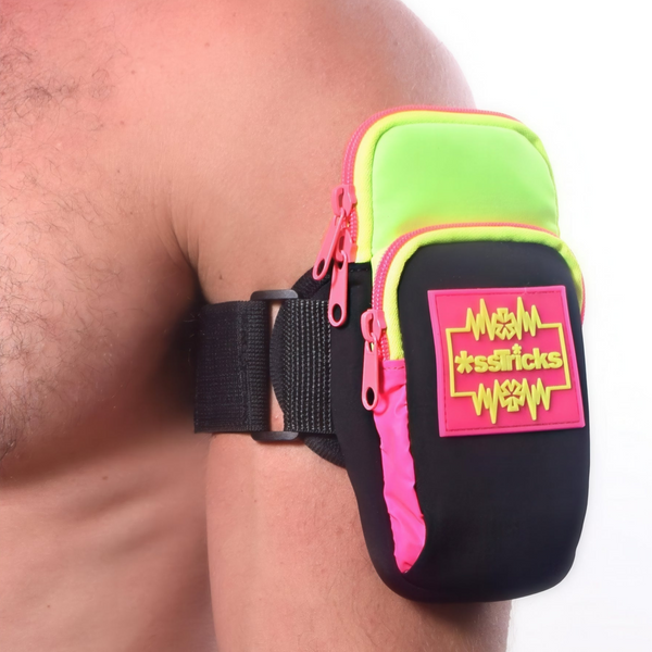 A person wears the Ekg Arm Bag in Neon by *ssTricks in green, pink, and black, featuring reflective side paneling, two zippered pockets, and a â*ssTricksâ patch on the upper arm. The background is plain white.