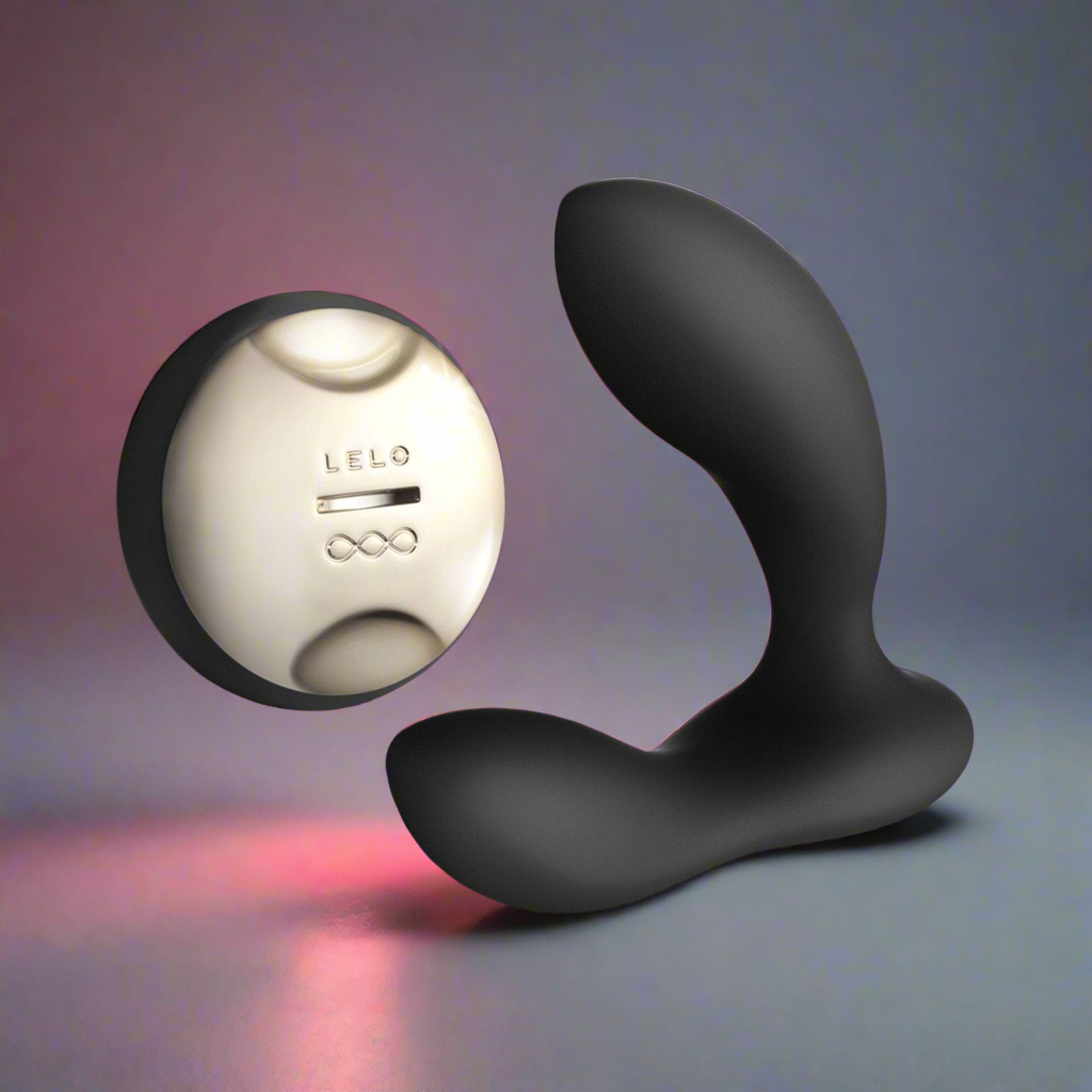 The LELO HUGO, a sleek black curved prostate massager, stands upright next to a round silver wireless remote labeled LELO. Both are placed on a smooth surface under soft gradient lighting.