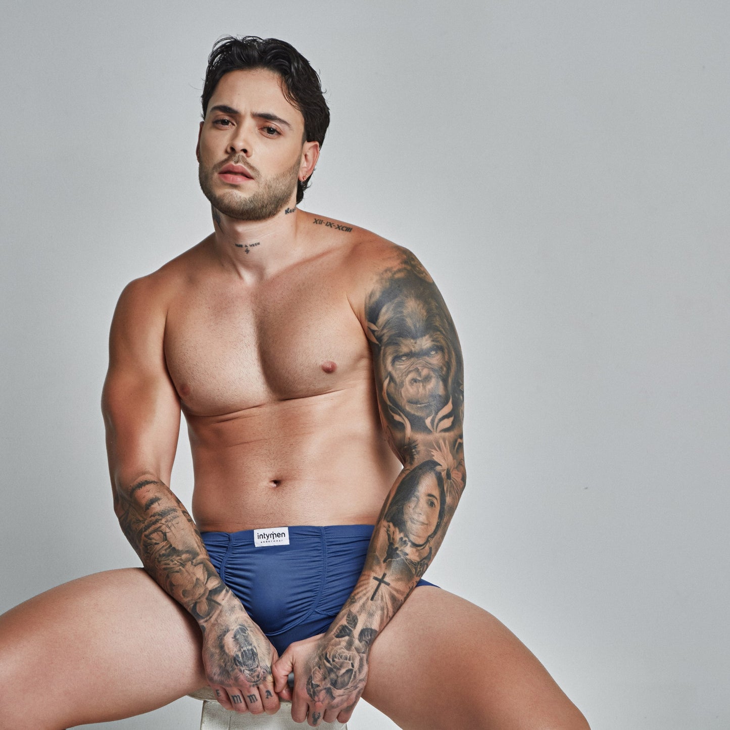 A tattooed man with short dark hair and a beard sits on a stool in Intymen Star Pouch Boxer Trunk Underwear, featuring soft, stretchy fabric and a spacious pouch for added comfort. He maintains a neutral expression against a plain gray background.