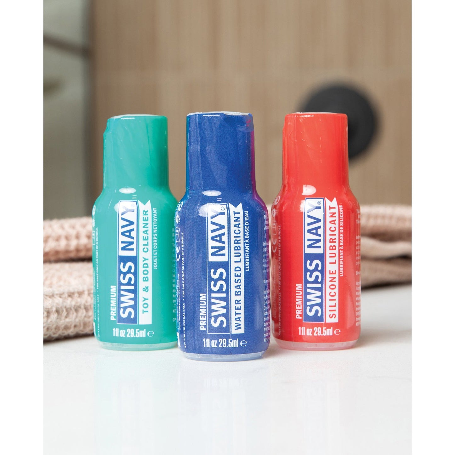 The Swiss Navy Essentials Variety Pack of 3, featuring green Toy & Body Cleaner, blue Water-Based Lubricant, and red Silicone Lubricant bottles, is elegantly arranged on a towel by Swiss Navy.