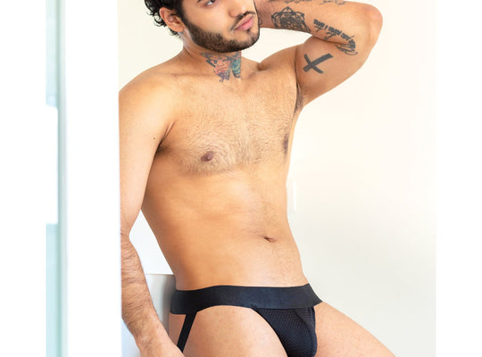 A tattooed man confidently poses in the 665 Stealth Jockstrap by 665 Inc, featuring a black design with a see-through mesh pouch, while resting one arm on his head against a neutral background.
