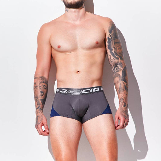 A tattooed individual stands against a plain background, highlighting Agacio Boxer Trunks Comfortable Underwear in gray and black. Their visible upper body and legs emphasize the contour pouch magic design by Agacio for ultimate comfort.