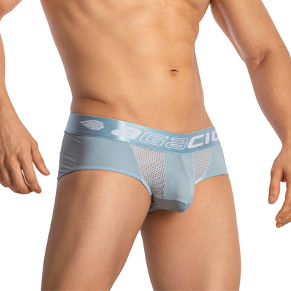 The Agacio Sheer Boxer Briefs in light blue have a wide waistband with white lettering and a small logo. The mesh front highlights the supportive pouch, while hands rest casually at their sides, focusing on the contoured lower torso design.