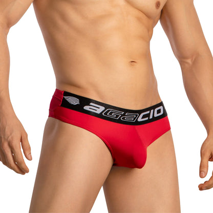 A man is shown wearing Agacio Thongs for Guys Sports Underwear, featuring a red fabric with a black waistband labeled Agacio. The sleek sporty design enhances his toned torso and arms, while the supportive contour pouch adds comfort against a crisp white background.