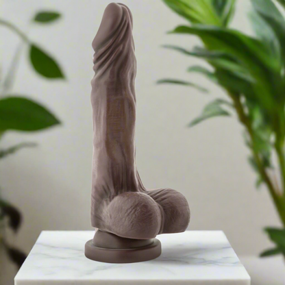 The Blush Novelties Dr. Skin - Mr. Skin Stud Muffin in chocolate, standing 6.25 inches tall, features a phthalate-free, realistic design resembling a human phallus, displayed upright on marble with a background of blurred green plants.