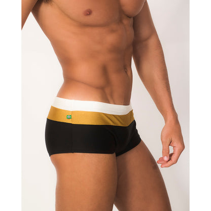 A model showcases the Belmont Trio Sunga by CA-RIO-CA Sunga Co., featuring handmade designer swim shorts in black and gold with a white waistband, highlighting their luxurious fabrics and striking design against a plain background.