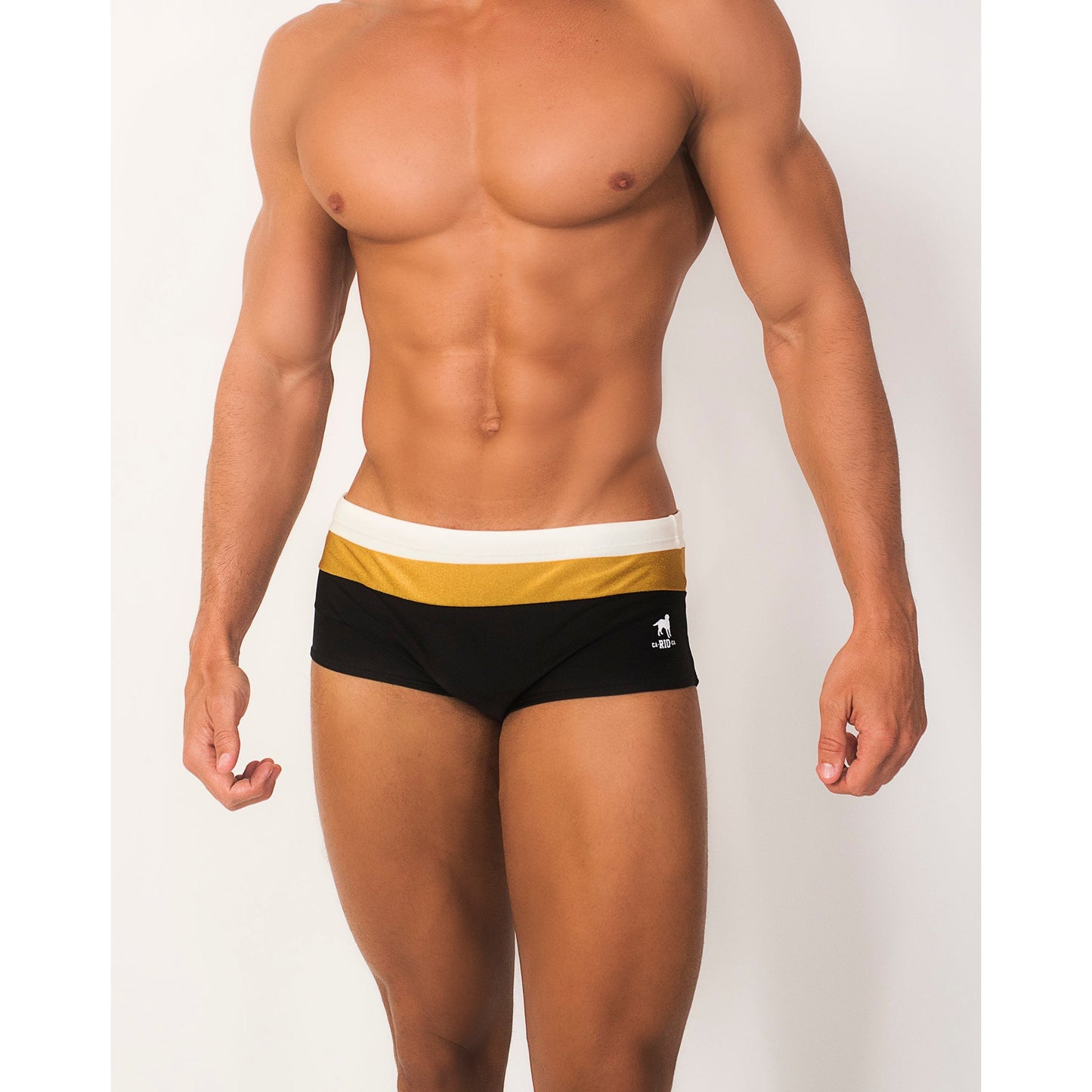 A muscular model wears the CA-RIO-CA Sunga Co. Belmont Trio Sunga, black and gold designer swim shorts with a luxurious fabric, featuring a wide white waistband and a small logo on the left. The model stands against a plain white background with their head not visible.