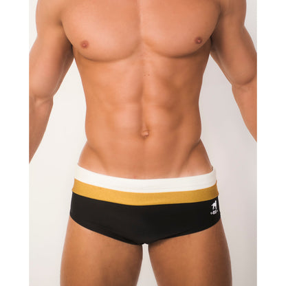 A male model displays CA-RIO-CA Sunga Co.s Belmont Trio Sunga, black and gold designer swim shorts with a white waistband featuring custom prints. The brand logo is on the right side of the waistband. Against a plain white background, his fit torso highlights the exquisite handmade detail.