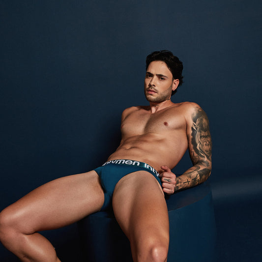 A tattooed man with dark hair leans casually against a dark blue backdrop, wearing Intymen Sky Jockstrap underwear. His serious expression exudes a mix of ease and sophistication as he rests slightly on a large, round object.