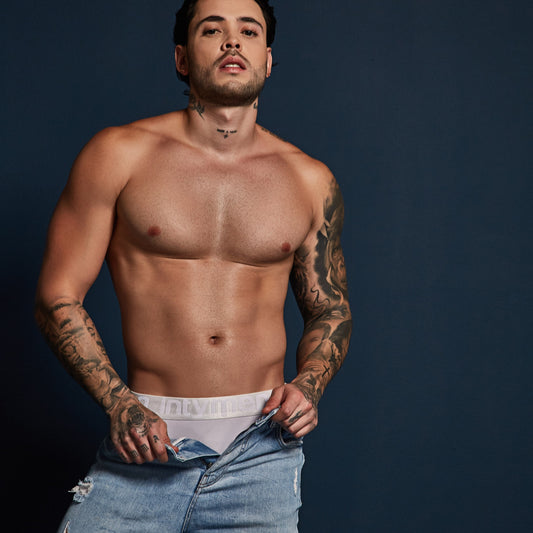 A tattooed man poses shirtless against a dark background, looking up. He wears light-washed denim jeans and an Intymen Energy Brief underneath, slightly pulling down the jeans to reveal the waistband, showcasing a figure-hugging fit.