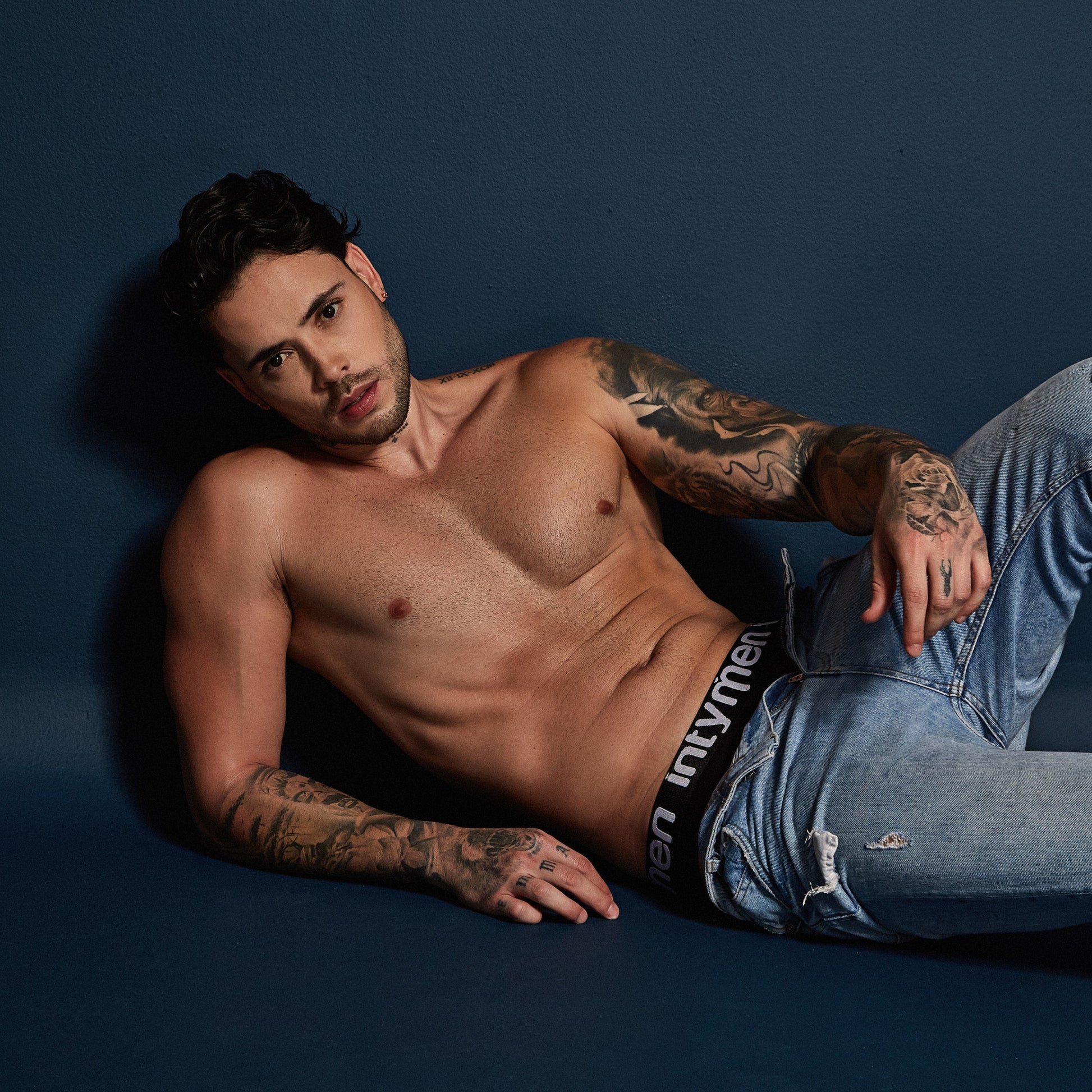 A tattooed shirtless man reclines on a dark blue surface, wearing jeans. An Intymen Sky Jockstrap Underwear for Men subtly peeks out, adding a luxurious touch. Gazing at the camera, he confidently displays his muscular physique and intricate tattoos.