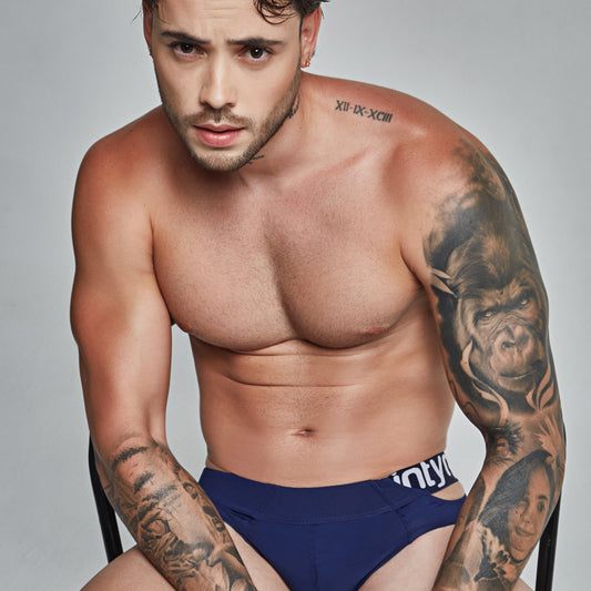 A tattooed man with short hair and a beard sits contemplatively on a chair, wearing navy blue Intymen True Brief Underwear. A Roman numeral tattoo adorns his shoulder against a plain background, highlighting the premium fabric blend.