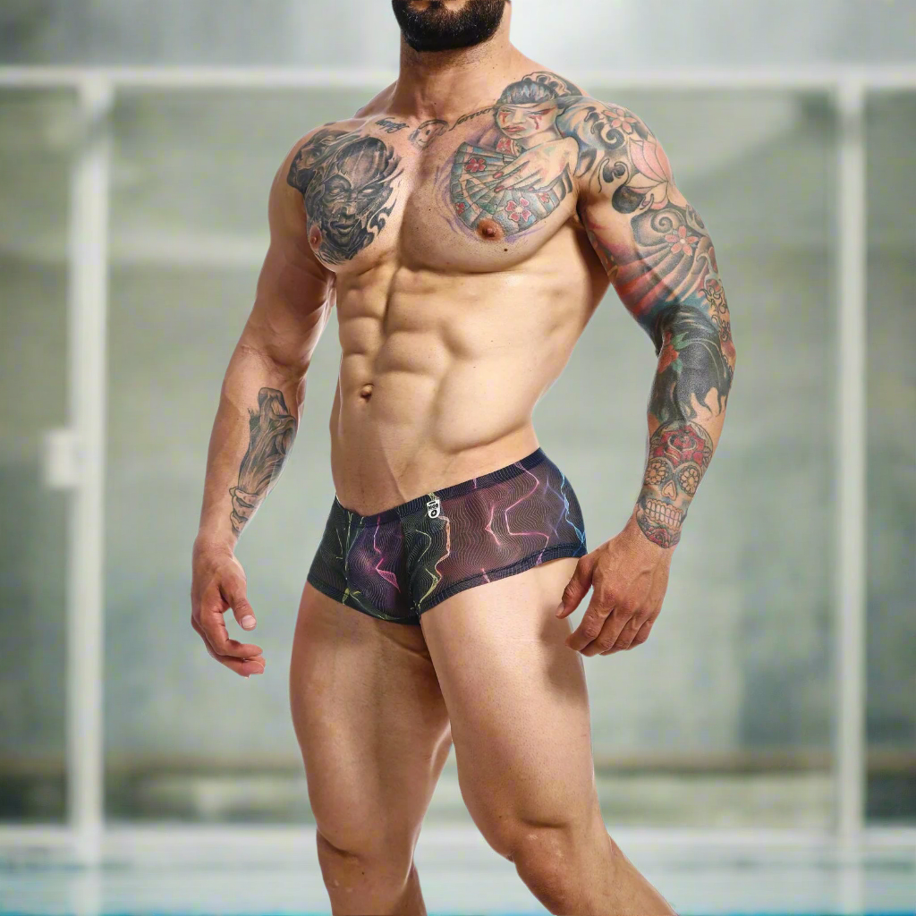 A muscular man stands confidently in Malebasics MOB Hip Hugger Boyshort Energy swim trunks, hugging his hips. His tattoo-adorned upper body and arms draw attention. The tastefully blurred background enhances the bold statement of this piece.