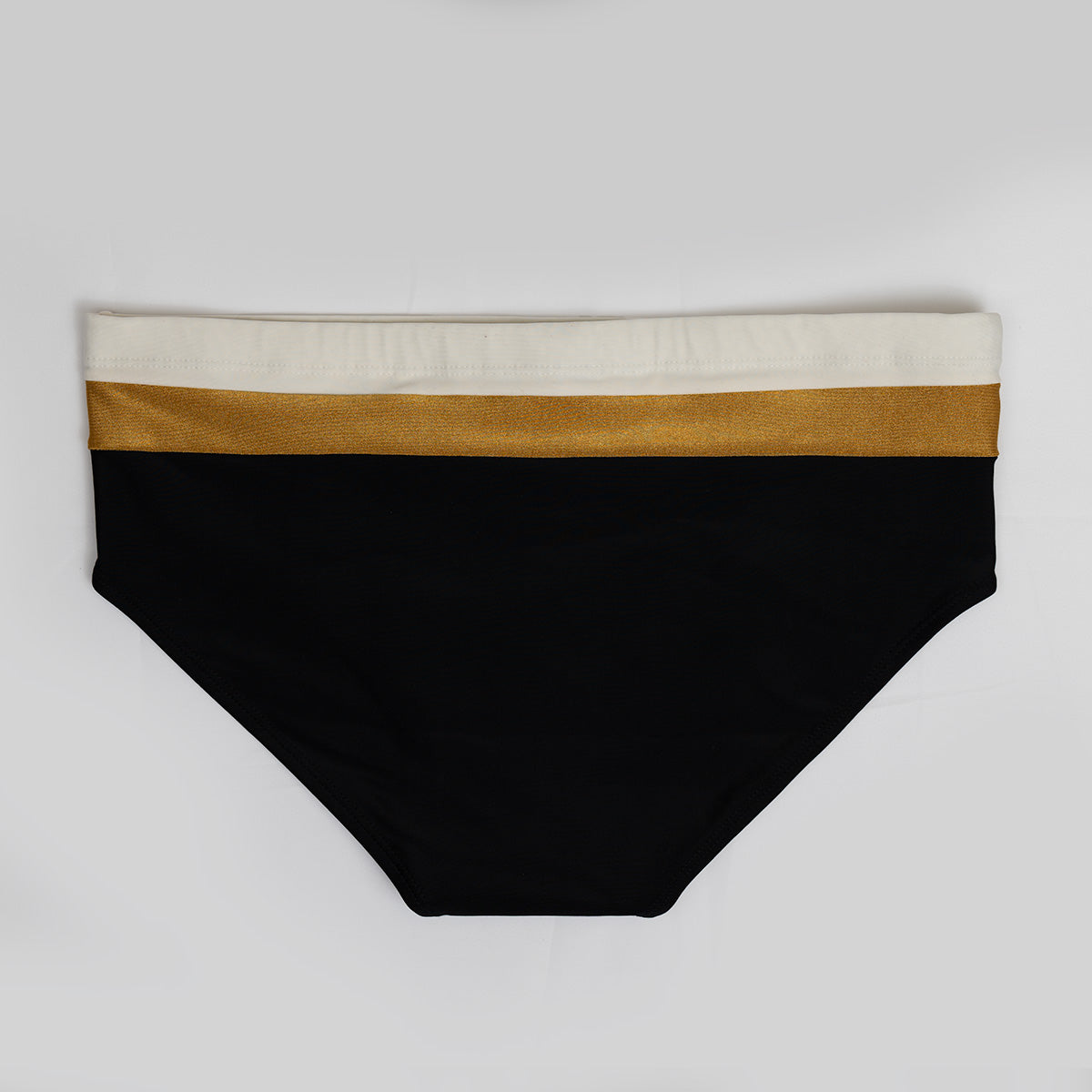 The Belmont Trio Sunga by CA-RIO-CA Sunga Co. features luxurious black bikini bottoms with a wide gold band and a narrower white band at the waist, all set against a plain gray background.