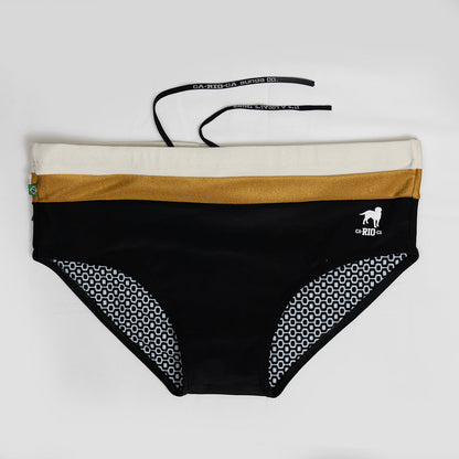 The Belmont Trio Sunga from CA-RIO-CA Sunga Co. is a luxurious mens swim brief with a white waistband, gold accent strip, and black body. It features black and white geometric edges, a small dog logo with Carioca text on the left, and functional drawstrings for added convenience.