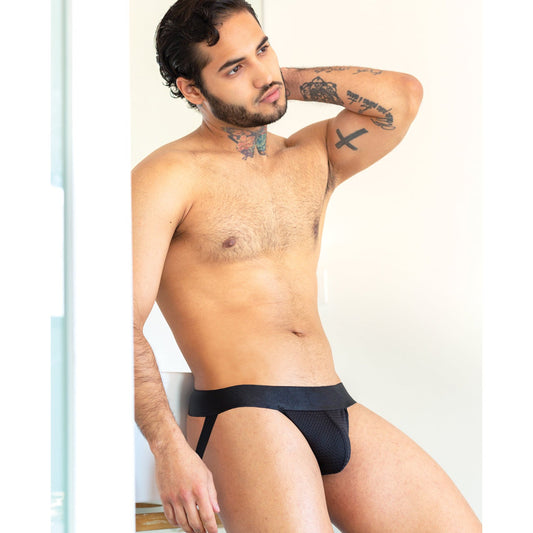 A tattooed man confidently poses in the 665 Stealth Jockstrap by 665 Inc, featuring a black design with a see-through mesh pouch, while resting one arm on his head against a neutral background.