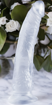 A translucent silicone sculpture, mimicking an organic form, stands on a marble surface, echoing the craftsmanship of Pipedreams Basix Rubber Works Slim Dong 7. In the background, there is a bouquet of green leaves and white roses.