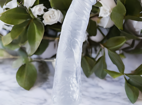 A translucent silicone sculpture, mimicking an organic form, stands on a marble surface, echoing the craftsmanship of Pipedreams Basix Rubber Works Slim Dong 7. In the background, there is a bouquet of green leaves and white roses.