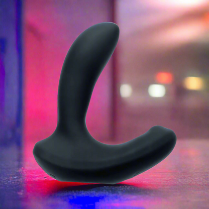 The VeDO Volt Rechargeable Prostate Vibe - Black stands upright on a reflective surface, its adjustable shaft highlighted against a backdrop of blurred, colorful lights that enhance the vibrant and moody atmosphere.