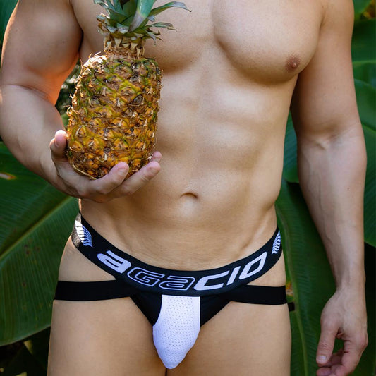 A shirtless person with a pineapple stands against large green leaves, wearing the Agacio Thriller Jockstrap featuring a distinctive white and black waistband, giving them an exotic appearance.