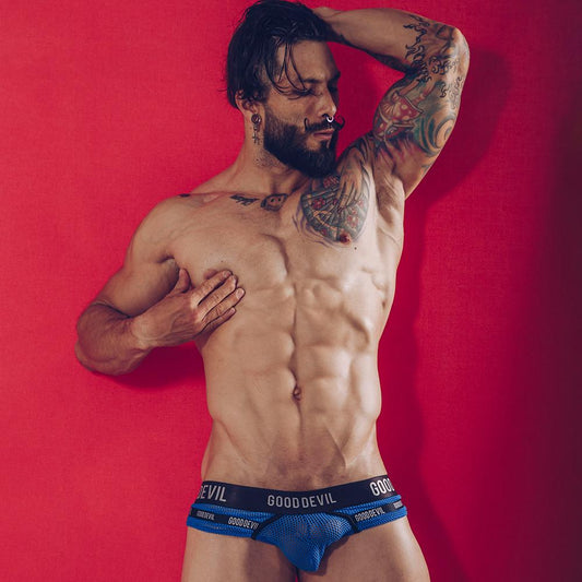 A muscular, tattooed man poses confidently against a red background, wearing Good Devils Show It All Briefs. The mesh fabric reveals his toned abs as he rests his hand on his head.