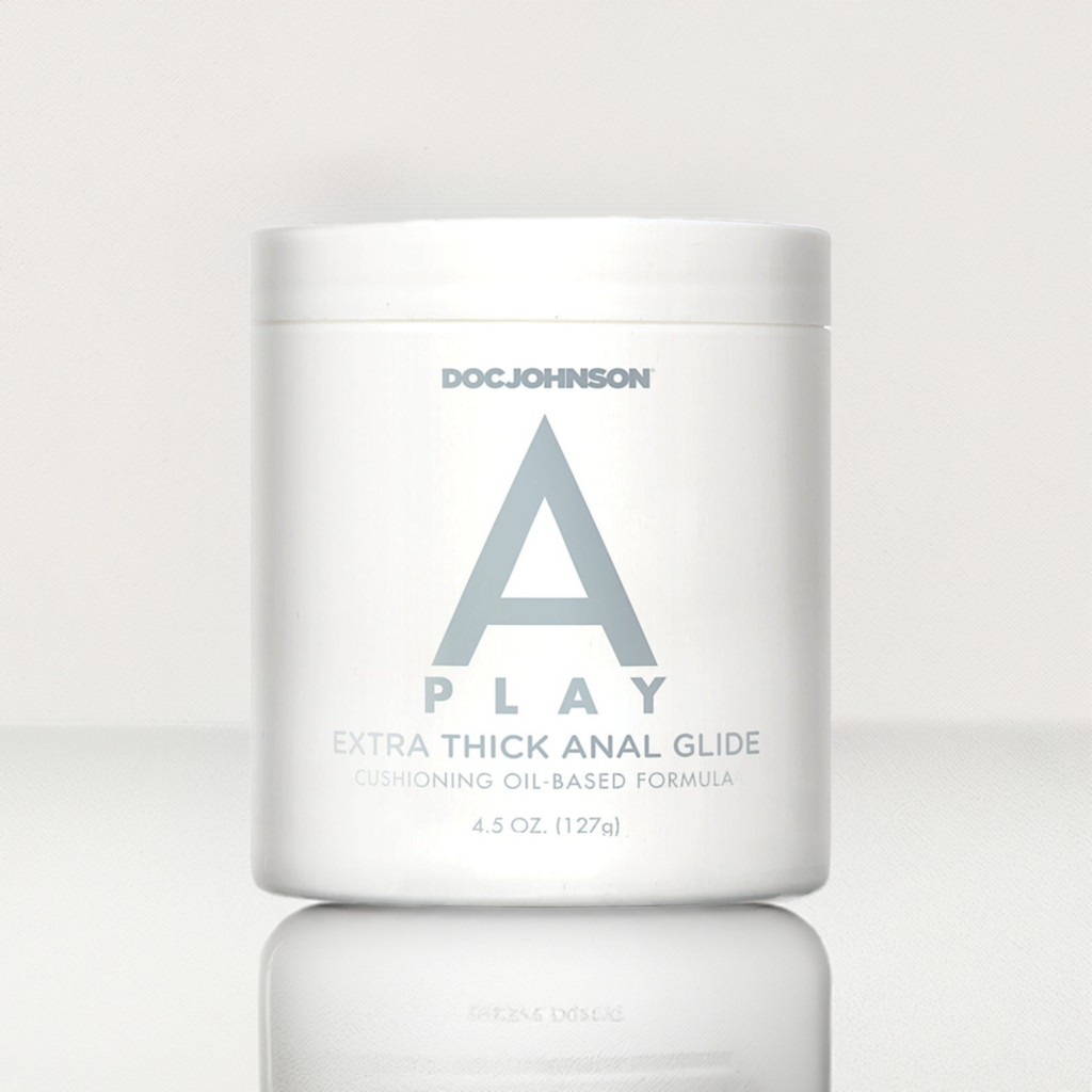 The image displays a white jar labeled A Play Extra Thick Anal Glide Cushioning Oil-Based Formula, featuring a large gray A. The 4.5 oz (127g) container rests on a reflective surface, emphasizing its sleek design and premium quality.