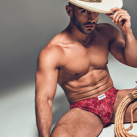 A shirtless man in Intymen Sexy Boy Paisley Bikini briefs sits on the ground, holding a cowboy hat. A coil of rope lies next to him, and the neutral background emphasizes both him and his attire.