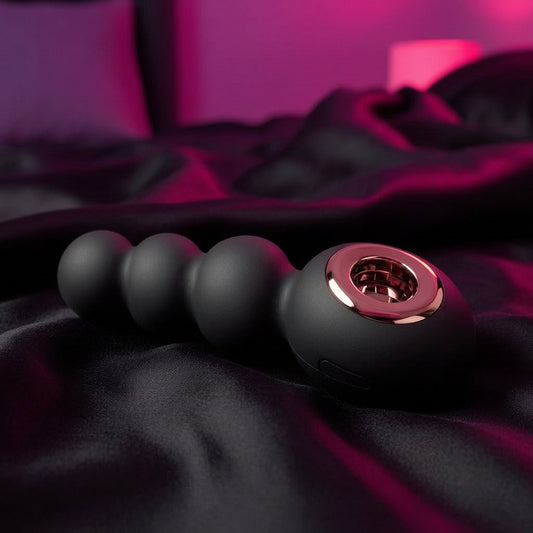 The Gender X Quiver Anal Ring Bead Vibe, crafted from medical-grade silicone with a rose gold accent, rests on black satin as soft pink lighting sets a sensual mood.