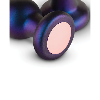 Close-up of a metallic object from the Hueman Comets Anal Trainer Butt Plug Set, showcasing a smooth, round base with a purple-blue gradient. The front has a circular pale pink area contrasting against its darker surface.