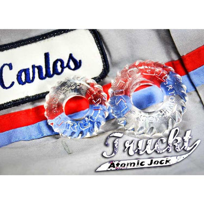 Close-up of two clear Skinflex TPR rings on a gray fabric with red and blue stripes, featuring a label that reads Carlos and brand identifiers Truckt and Atomic Jock, emphasizing the Oxballs TruckT Cock & Ball Ring - Pack of 2.